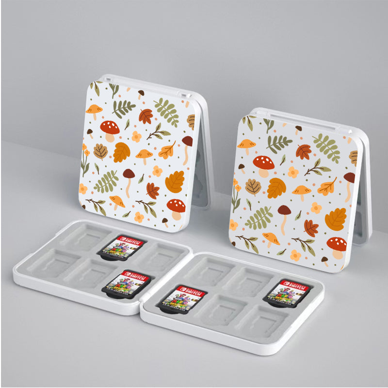 Two open Cozy Autumn Game Card Boxes for Nintendo Switch by petimint, each holding four small memory cards, are displayed on a light grey surface. The lids feature a playful autumn-themed design with leaves, mushrooms, and acorns in shades of orange, brown, and green. These cases boast magnetic closures for secured storage.
