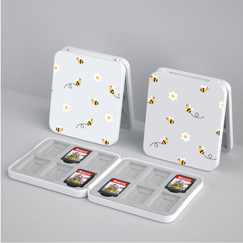 Two white "Little Bee Game Card Boxes for Nintendo Switch" by petimint, featuring square designs with bee and flower patterns on their covers, are open to display slots for storing game cards and micro SD cards. One case reveals Mario Kart and Super Mario game cards inside. Perfect for the cozy gamer, the cases sit on a light gray surface.