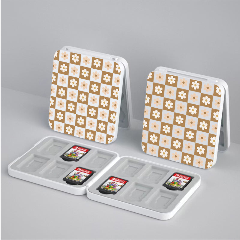 Two small, square silicone cases named "Cozy Checkered Flowers Game Card Box for Nintendo Switch" by petimint, feature floral and checkered patterns on their exteriors. Both cases are open, revealing four game card slots inside. Two game cards are placed in slots in the left case, while the right case is empty. The background is a plain surface.
