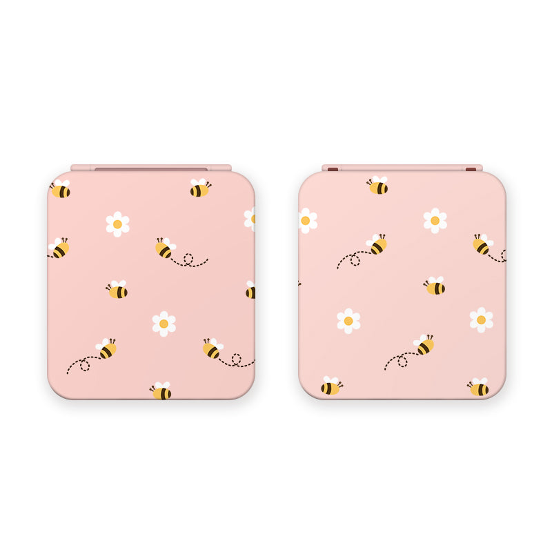 Two pink game card boxes from petimint are adorned with a whimsical design featuring small bees and white flowers. The bees, characterized by their black stripes, are depicted in various flying positions with dotted flight paths. Perfect for the cozygamer, the boxes showcase their charming exterior when closed.