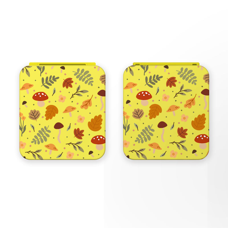 Two square yellow compact mirrors with an autumn-themed design featuring mushrooms, leaves, and ferns in various shades of brown, orange, and green are displayed side by side against a white background. Each mirror features a convenient magnetic closure.