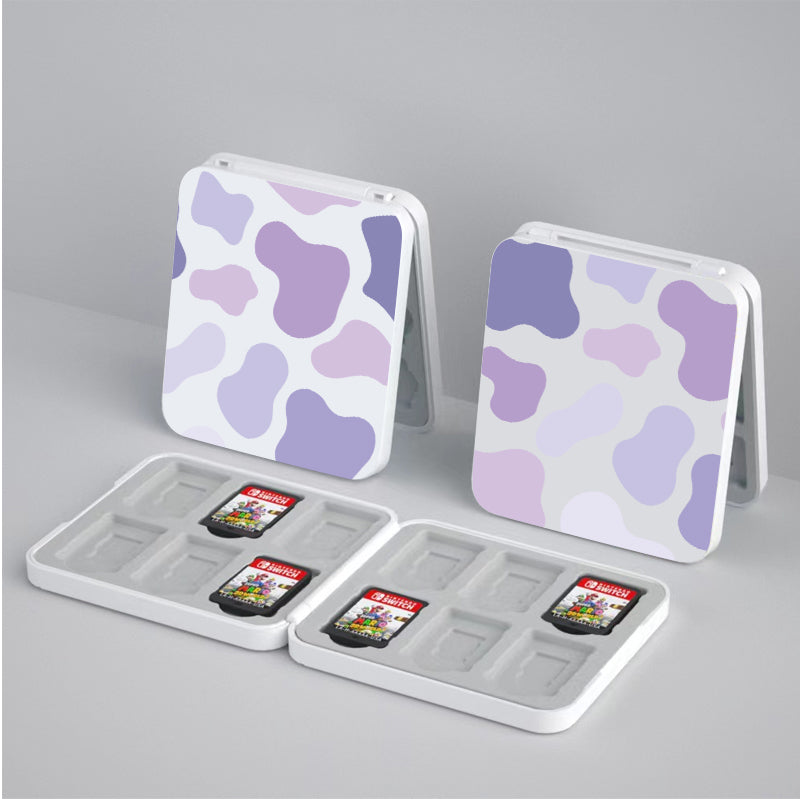 Two open Lilac Cow Game Card Boxes for Nintendo Switch by petimint with purple and lavender cow-print exteriors stand side by side. Each box contains several game cards, some of which have visible red labels. This gamer-friendly setup rests on a plain light gray surface, showcasing both style and function in your gaming attire.