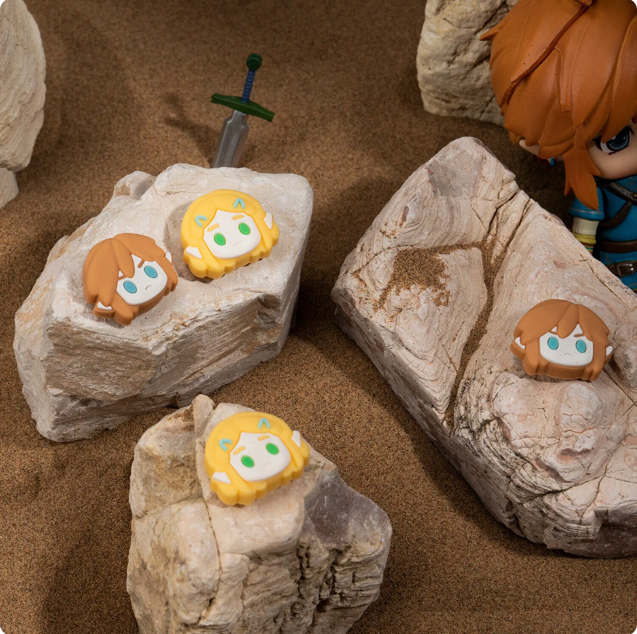 In a sandy environment, small, stylized figurines with distinct hair colors and simplistic facial features are placed on rocks. A larger figurine, standing partially visible on the right and holding a Nintendo Switch equipped with Zelda 3 Thumb Grips by petimint, adds to the scene while a sword stands embedded vertically in the background.