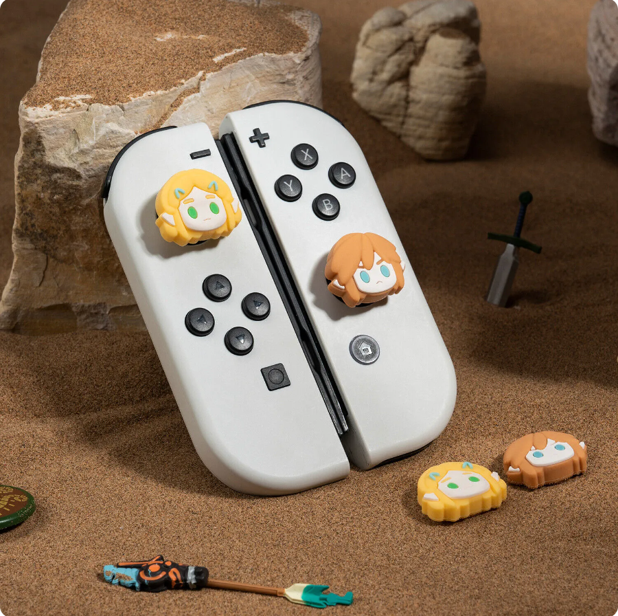 Two white Joy-Con controllers for the Nintendo Switch are adorned with petimint's Zelda 3 Thumb Grips, which feature character-themed designs with small, stylized faces. These controllers are displayed in a sandy environment complete with rocks and miniature props, including a toy sword and figure. Perfect for any gamer, the controllers lean against a rock.