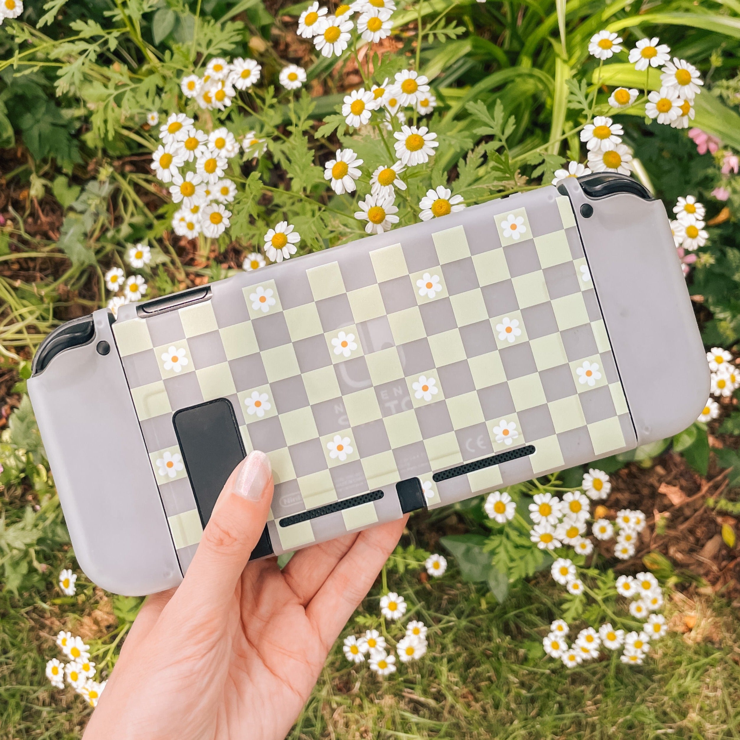 A hand holding a Nintendo Switch encased in the petimint Checkered Flowers Case, which features small daisy patterns on a green and white checkered background. The scene is set against lush green foliage and clusters of blooming white daisies, creating an idyllic setting for any cozygamer.