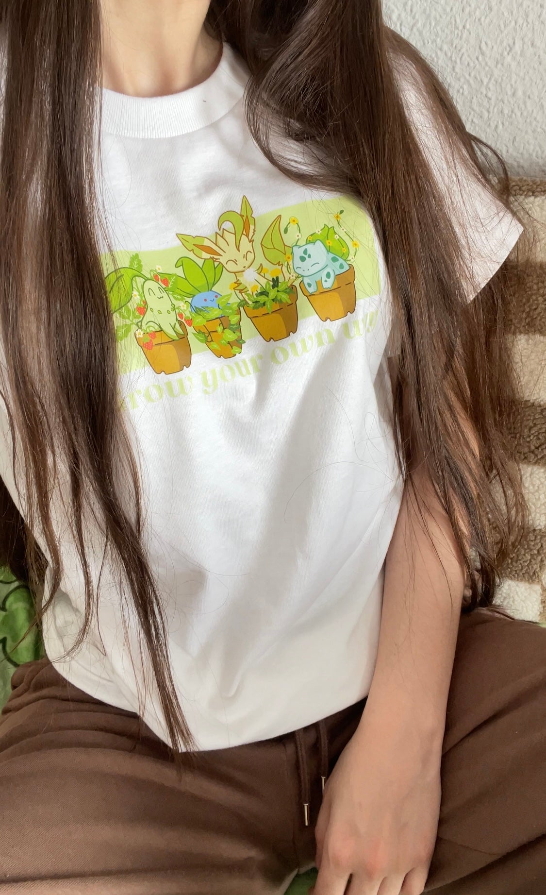 A person with long brown hair is sitting down, wearing a white T-shirt featuring cartoon illustrations of plant-themed characters in pots. The text on the shirt reads, "Grow your own way." The person, an avid gamer, is also wearing brown pants and has a Nintendo Switch by their side.