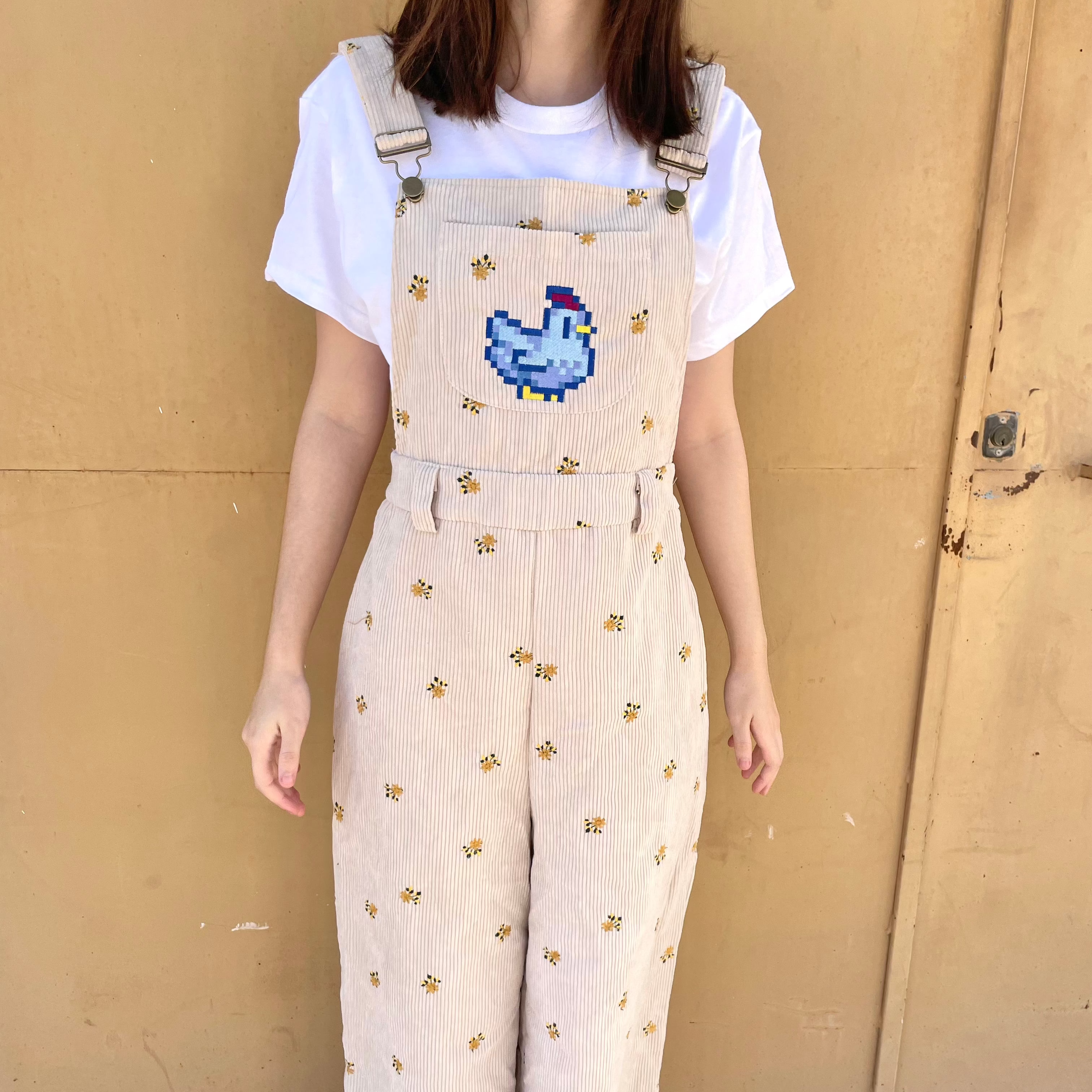 A person stands against a beige wall, wearing a white T-shirt under the petimint Stardew Chicken Floral Overalls Long Pants. The light beige corduroy overalls feature a pixelated blue bird design on the front bib and small floral patterns spread throughout, giving off major cozygamer vibes. The person's head is not visible in the image.