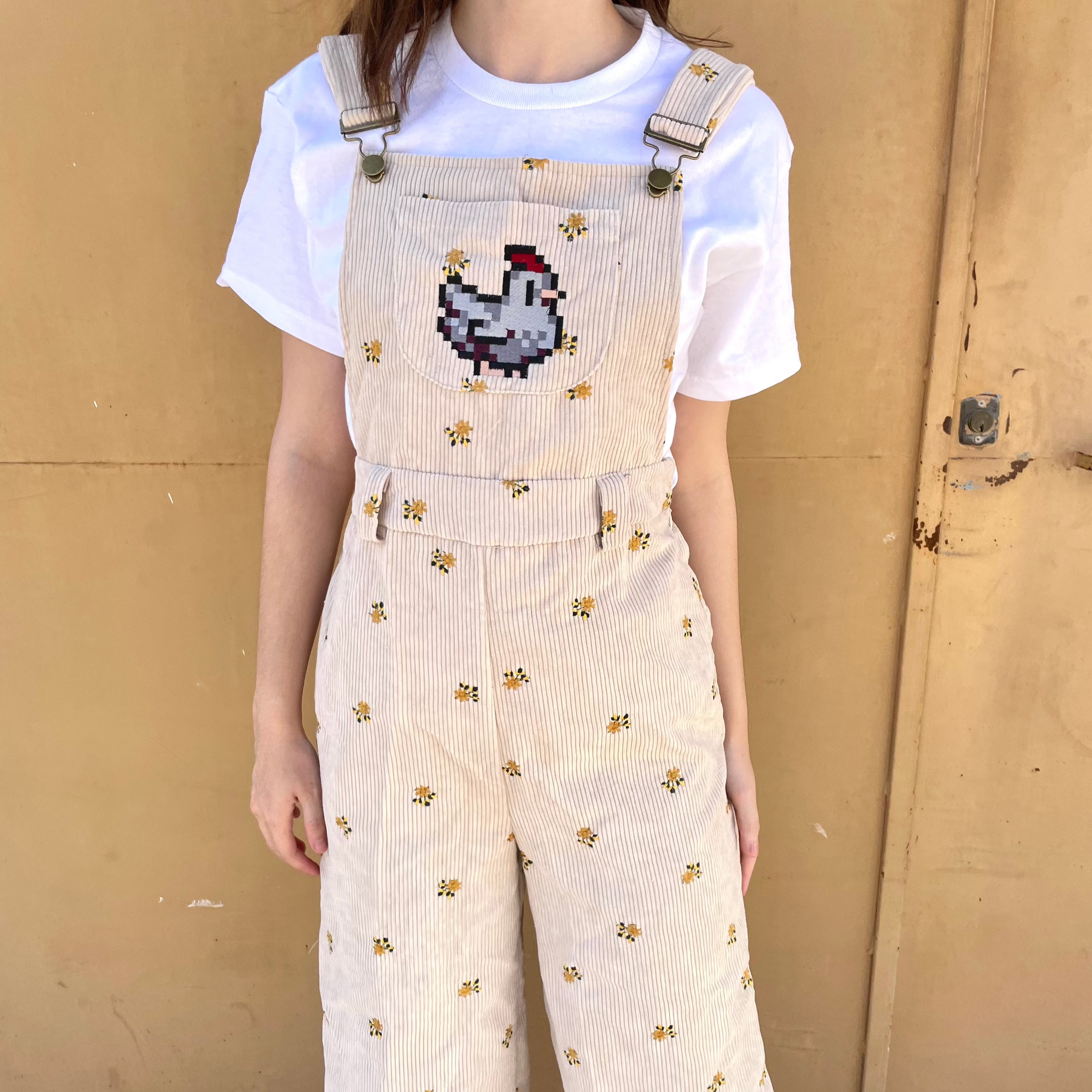 A person is standing against a beige wall wearing the petimint Stardew Chicken Floral Overalls Long Pants, featuring a small yellow floral pattern. A pixelated chicken, inspired by Stardew Valley, is prominently embroidered on the front of the overalls. They are also wearing a plain white t-shirt underneath.