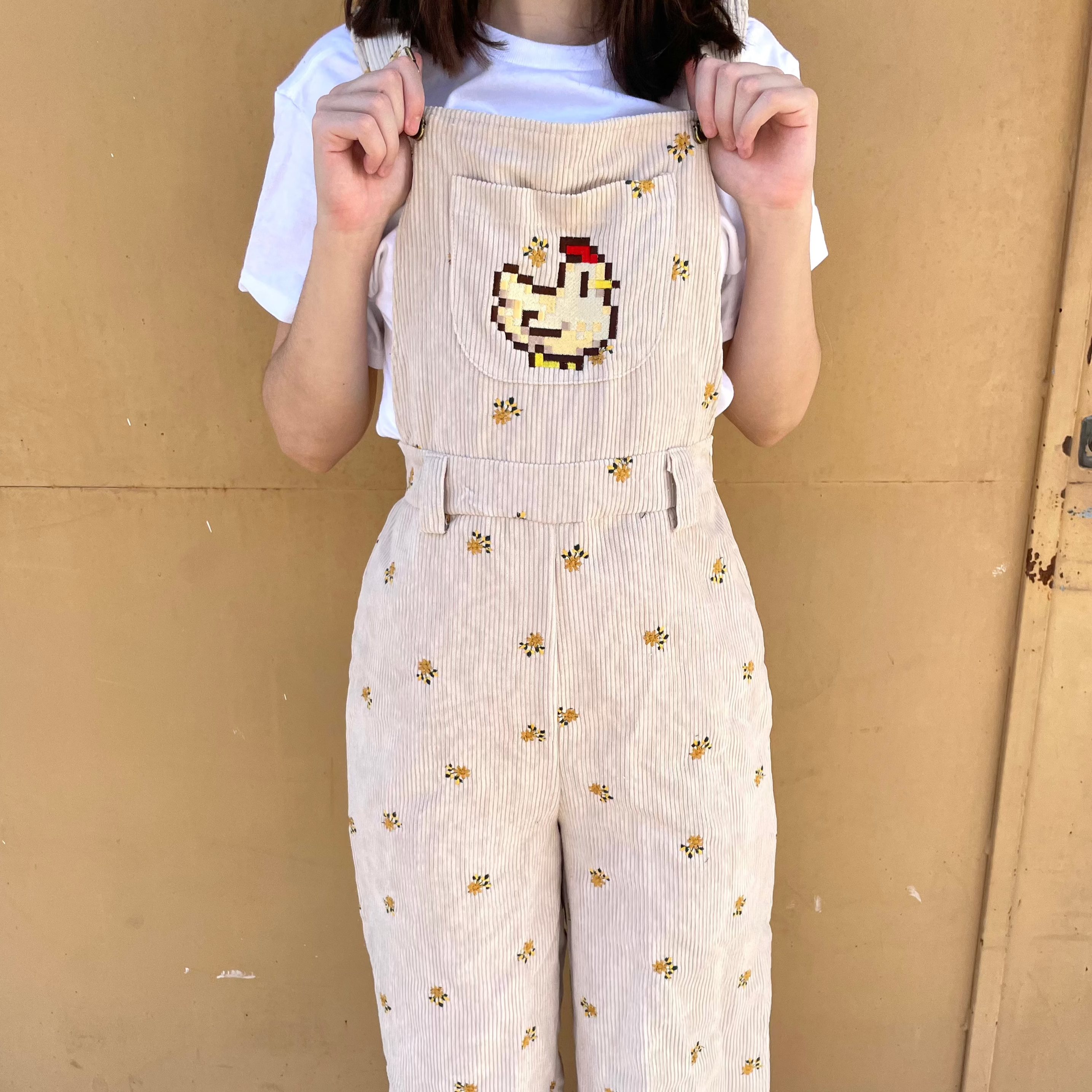 A person standing against a beige wall is wearing the 'Stardew Chicken Floral Overalls Long Pants' by petimint, featuring light-colored fabric adorned with small yellow flowers and an embroidered design of a chicken building snowballs on the front bib. The individual, possibly a cozygamer, pairs this outfit with a white T-shirt underneath.