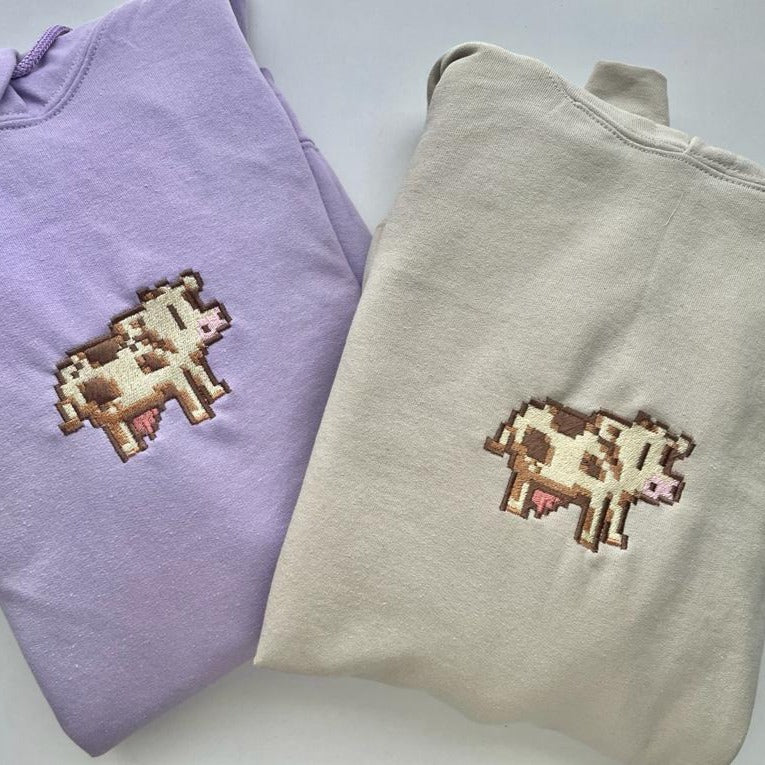 Two folded Stardew Cow Hoodie Apparel sweatshirts from petimint are displayed side by side. The sweatshirt on the left is light purple, while the one on the right is cream-colored. Both feature a pixelated cow design embroidered on the front, making them ideal for any cozygamer wanting to stay warm while playing on their Nintendo Switch.