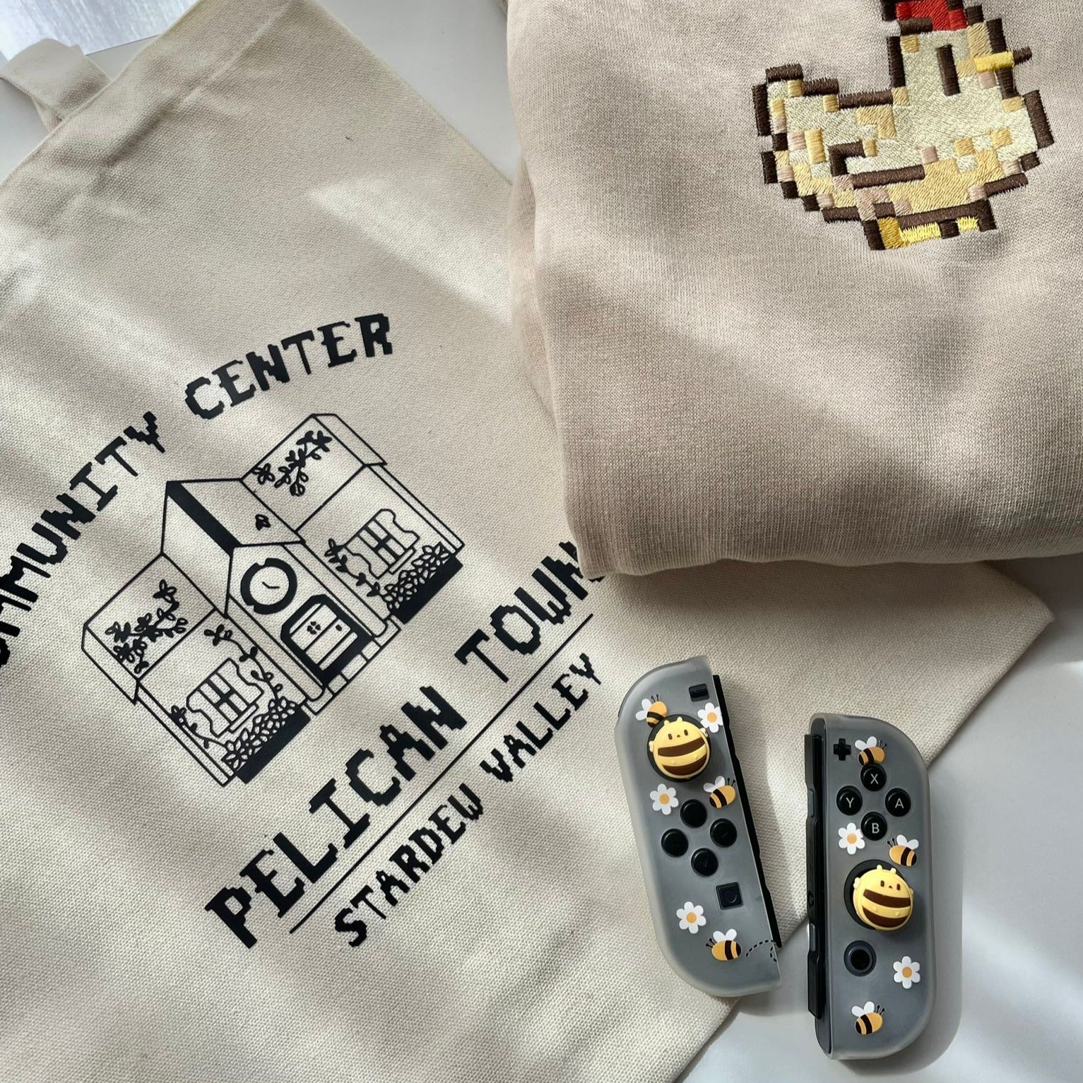 A petimint "Pelican Town Tote Bag," a cream-colored sweater featuring a pixel art chicken, and a pair of gaming controllers adorned with animal-themed thumb grips—all displayed on a flat surface. Perfect for any gamer!