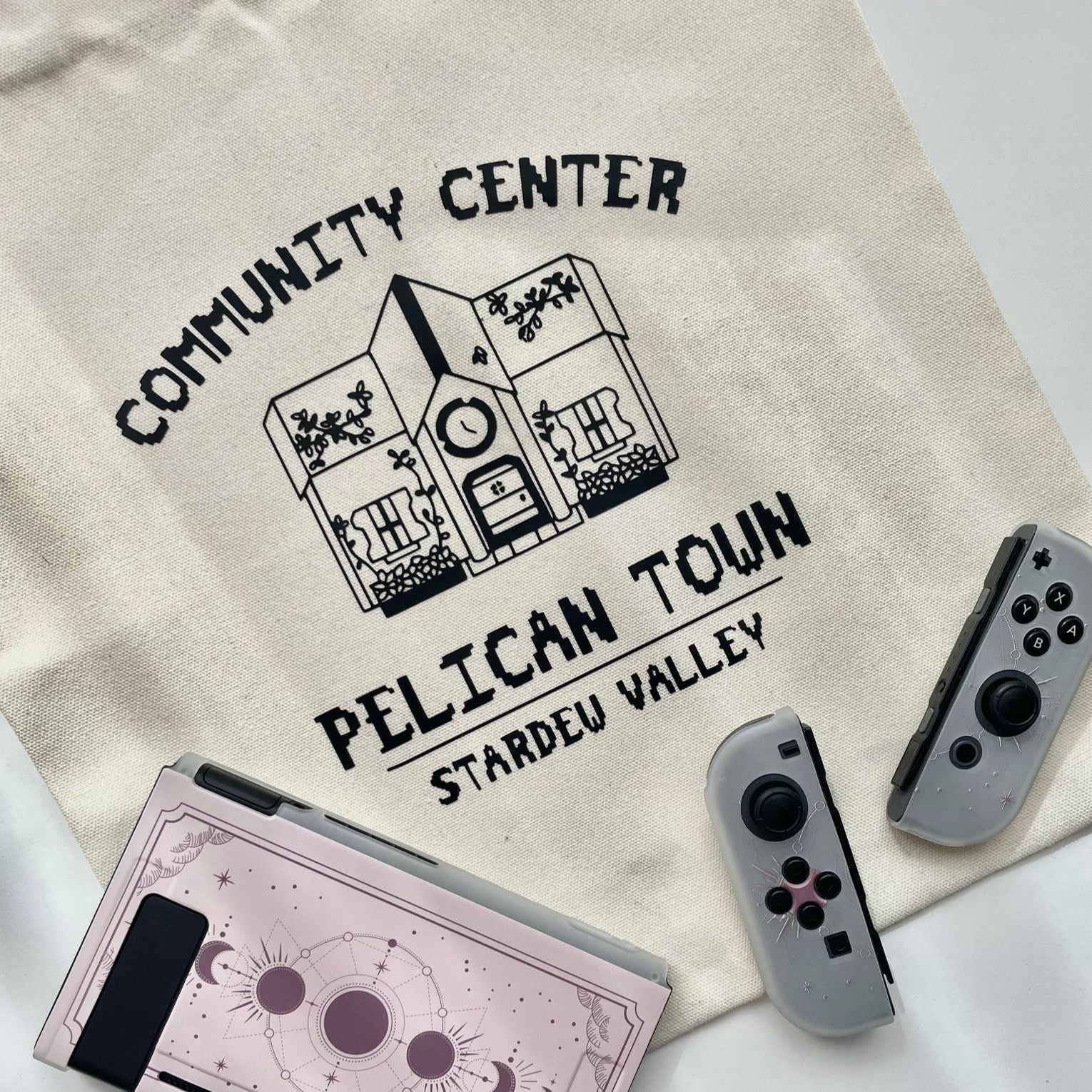A Pelican Town Tote Bag from petimint, featuring a black print of a building labeled "Community Center Pelican Town Stardew Valley," lies on a surface. Nearby, there is a Nintendo Switch console with a pink custom cover and two detached Joy-Con controllers, one black and one gray. Perfect for the cozy gamer who loves unique apparel.