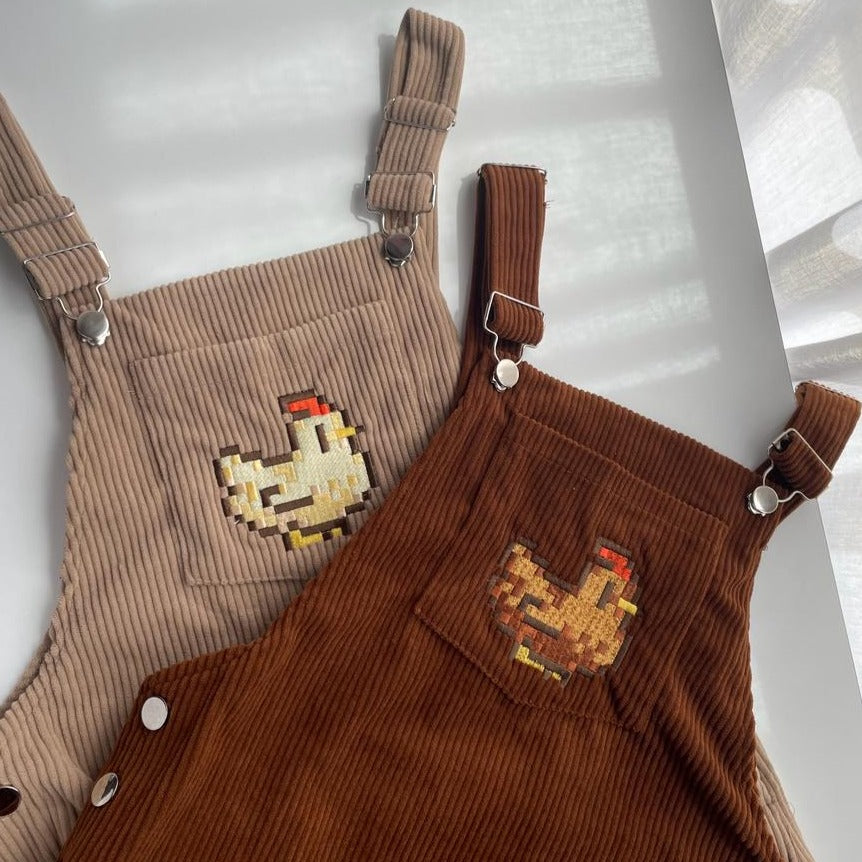 Two detailed corduroy overalls with charming chicken designs are laid out on a white surface. The left pair is beige and features a yellow and red chicken embroidered on the front pocket. The right pair, known as the Stardew Brown Chicken Overalls Dress by petimint, is brown with a brown and yellow chicken embroidered on its front pocket. Both items make delightful additions to any child's apparel collection.