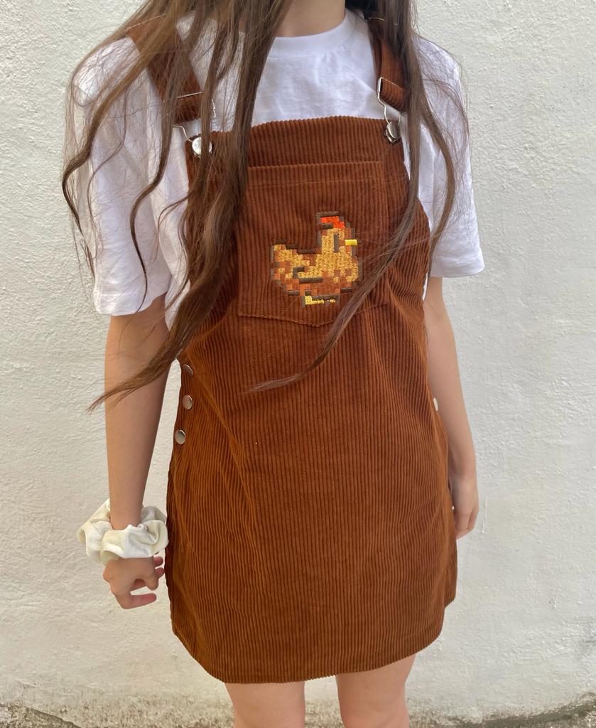 A person with long hair is wearing the petimint Stardew Brown Chicken Overalls Dress featuring a cross-stitched chicken design on the front pocket, paired with a white short-sleeve shirt. This cozygamer has a scrunchie on their left wrist and is standing against a plain white wall.