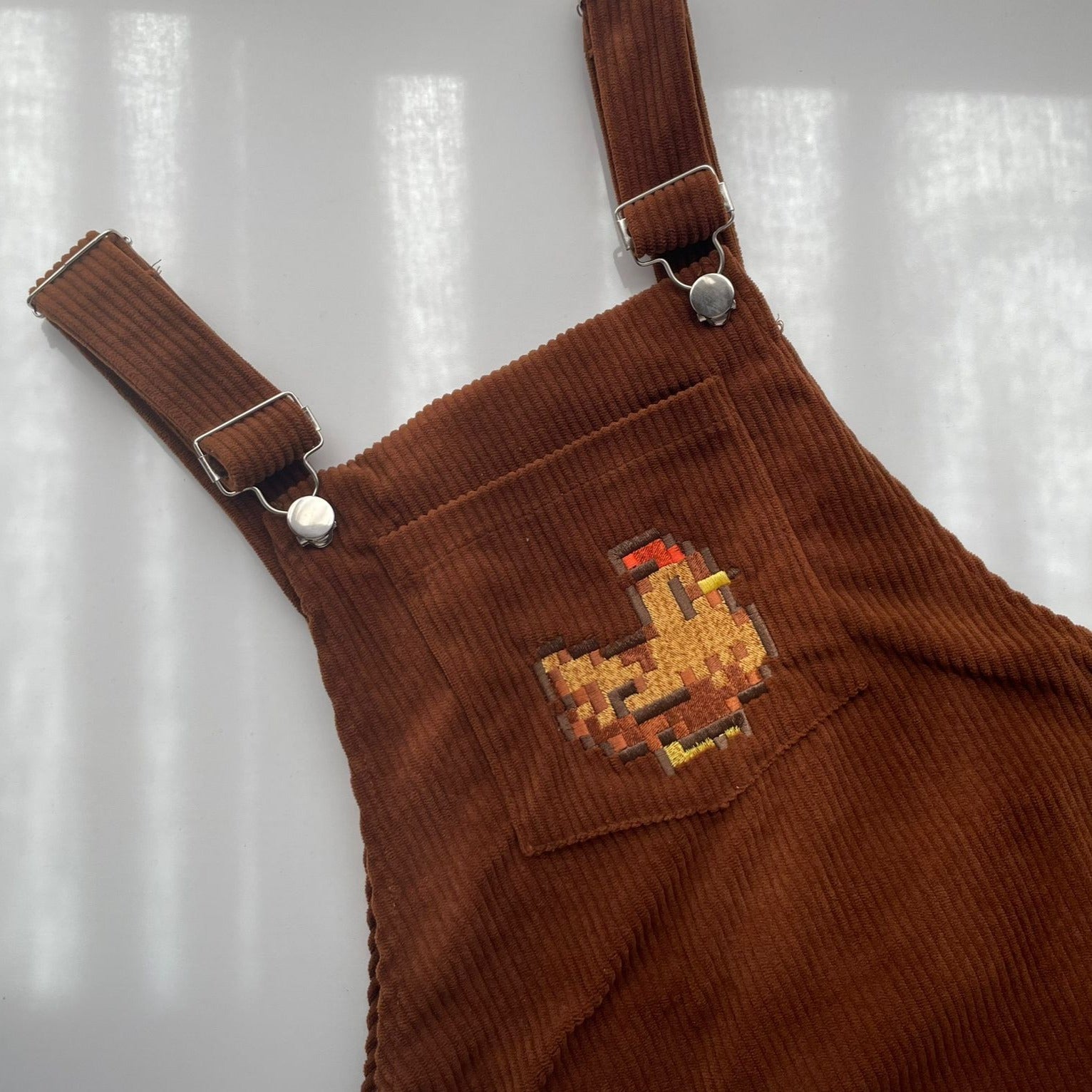 A close-up of the petimint Stardew Brown Chicken Overalls Dress showcases a small embroidered chicken on the chest pocket. The dress features strap buckles with silver buttons, and in the softly diffused light of the background, a Nintendo Switch can be casually spotted nearby.