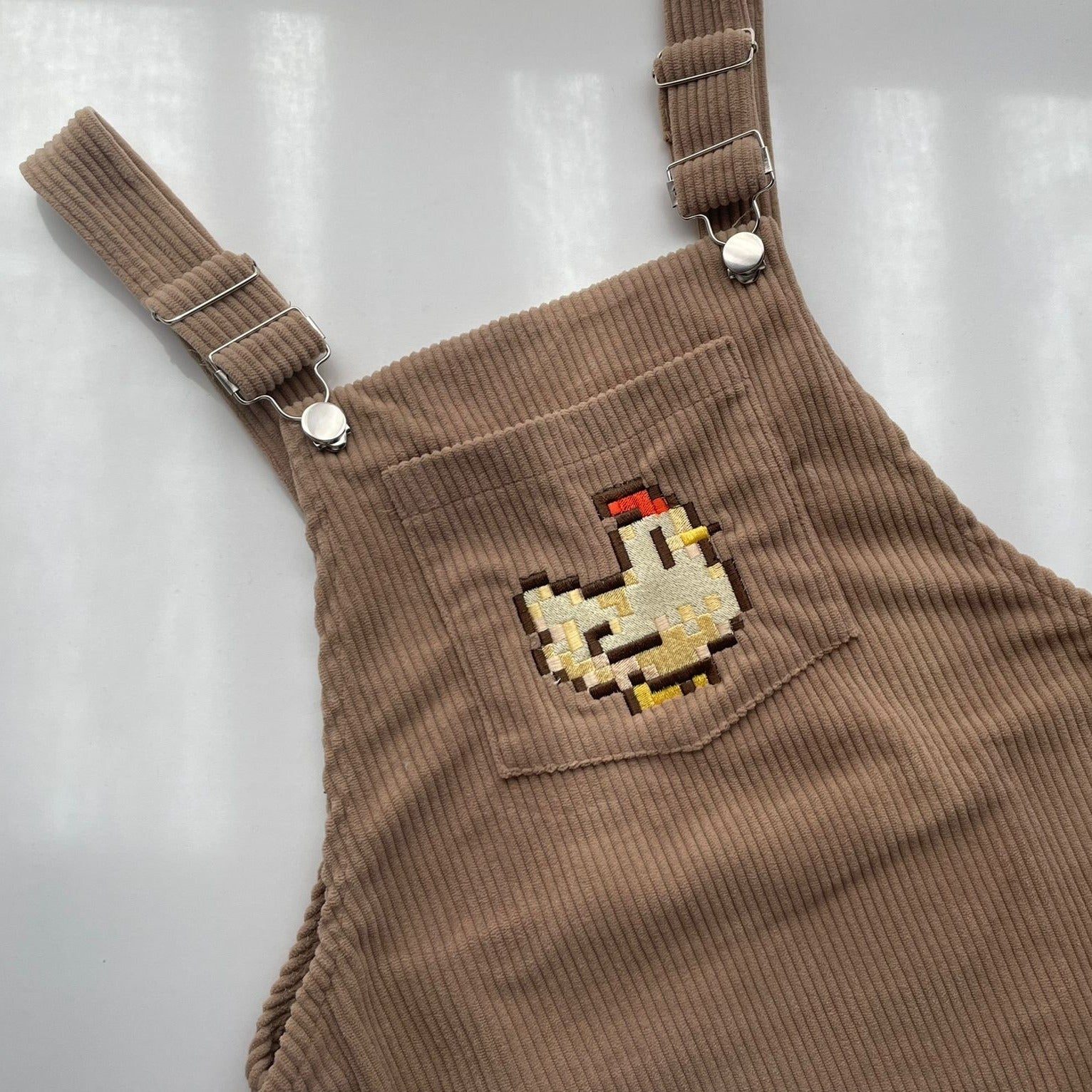 A close-up of the **petimint Stardew Chicken Overalls Dress** showcases a beige corduroy overall dress with adjustable straps and metal buckle closures. The front pocket boasts an embroidered pixel art design of a white chicken with a red comb and yellow beak, adding a playful, gaming-inspired touch to your apparel.