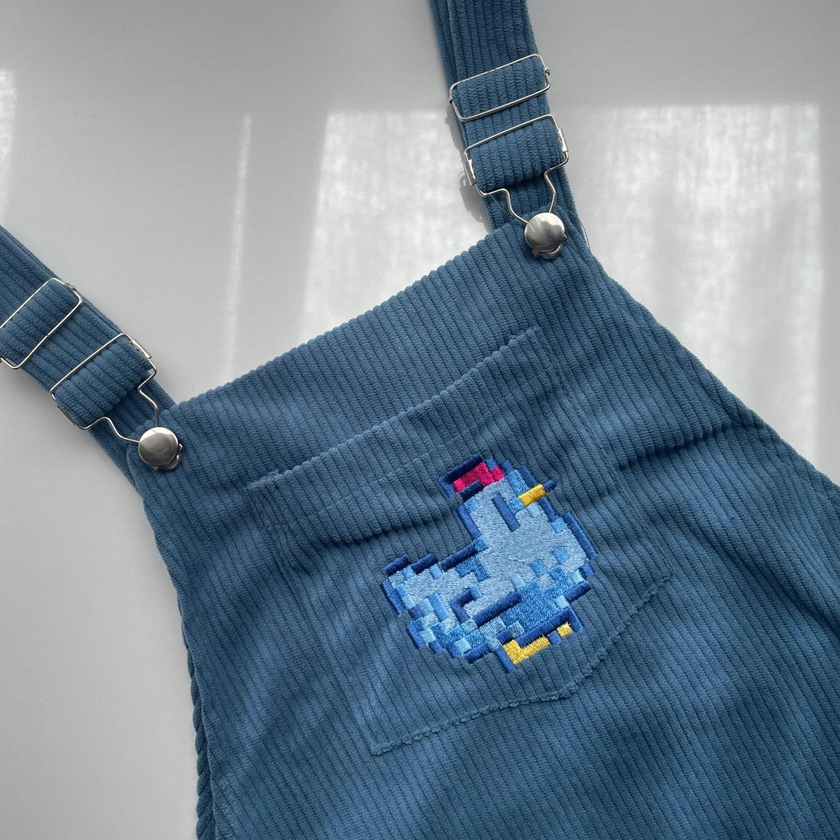 The Stardew Blue Chicken Overalls Dress by petimint is displayed, featuring adjustable metal buckles and a front chest pocket with a small embroidered pixelated chicken in shades of blue, yellow, and red. This comfortable piece is perfect for any cozygamer wanting to add a touch of gaming flair to their wardrobe.
