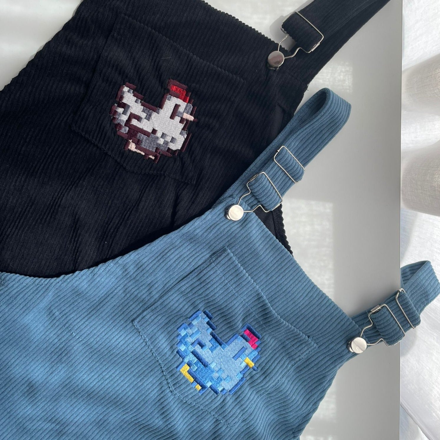 Two pairs of Stardew Blue Chicken Overalls Dress, crafted by petimint, are laid on a flat surface. One pair is black with a red and white pixelated chicken design on the chest pocket, while the other is blue featuring a blue and yellow chicken. Ideal apparel for any cozygamer, both overalls have silver buckles at the straps.