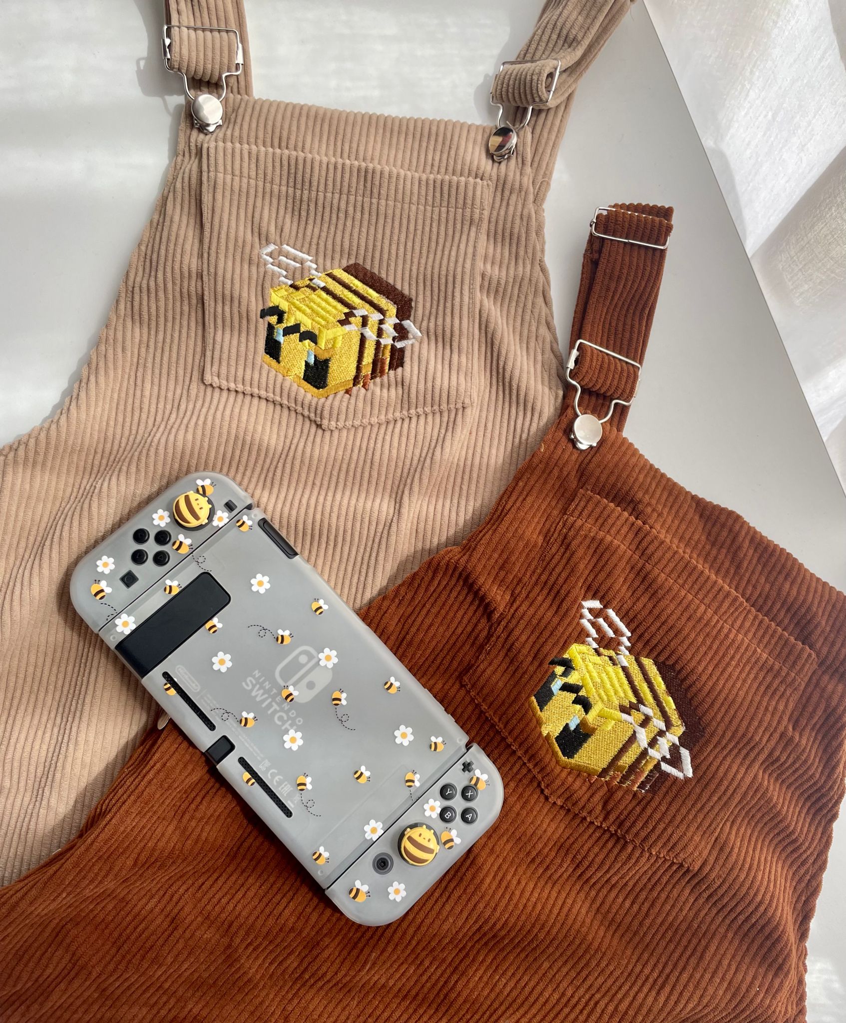 Two pairs of Bee Minecraft Overalls Dresses by Petimint, one in beige and the other in rust-colored, each adorned with an embroidered bee hive. In front of the dresses is a grey Nintendo Switch decorated with bee and flower stickers. The scene, ideal for a cozy gamer, is beautifully illuminated by natural daylight.