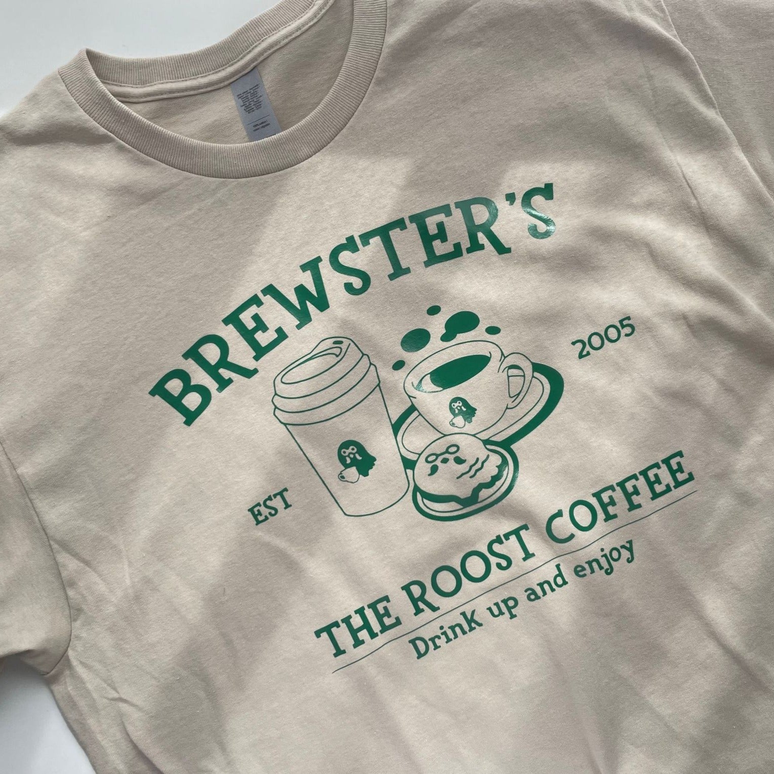 The Roost Coffee T-Shirt by petimint is a beige t-shirt with green text and graphics, featuring the words "BREWSTER'S, EST 2005, THE ROOST COFFEE, Drink up and enjoy." The graphics showcase a to-go coffee cup, a steaming mug, and a plate with a pastry—making it an ideal piece of apparel for any gamer inspired by Stardew Valley.
