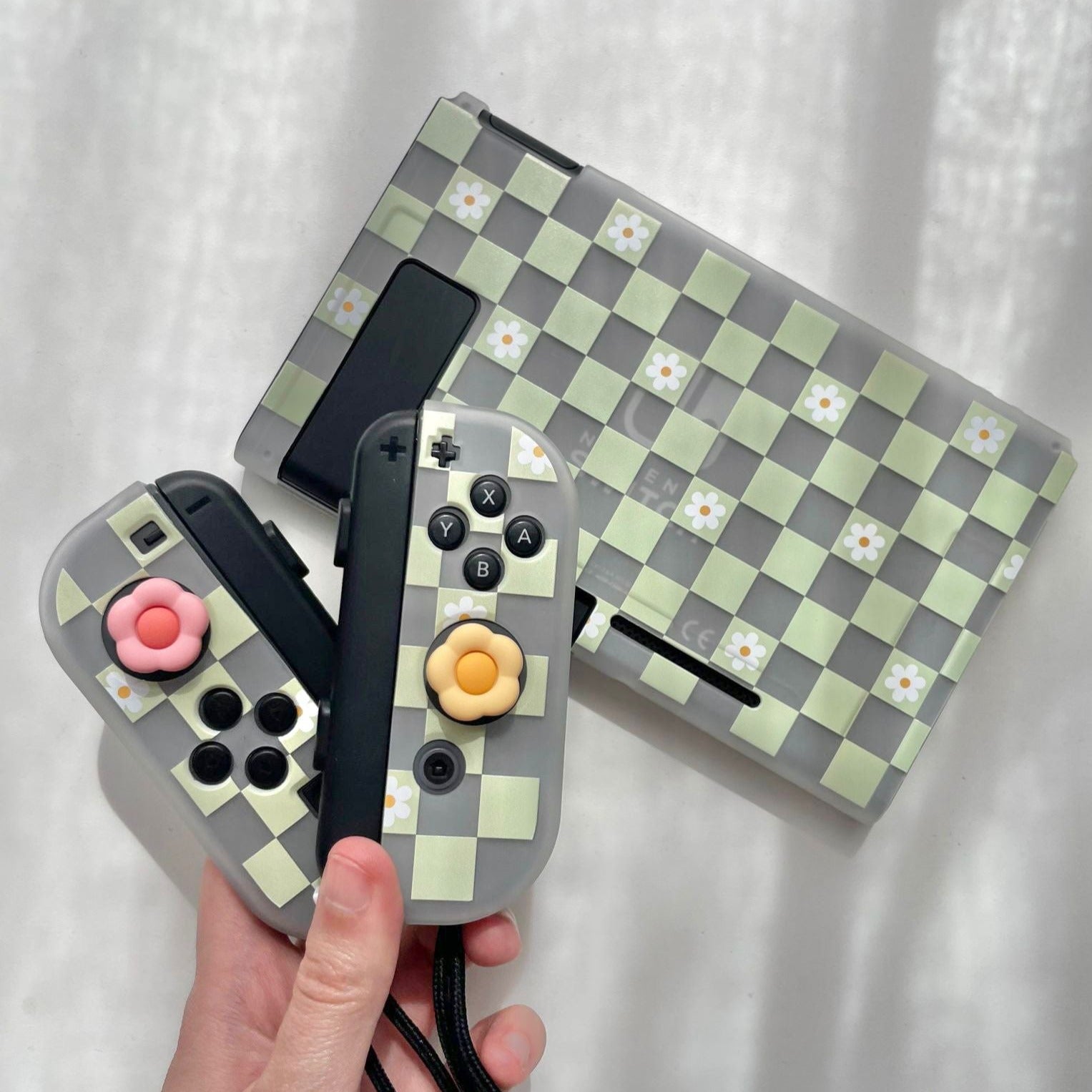 A hand holds a pair of custom Nintendo Switch controllers featuring a checkered green and white pattern with decorative flower buttons. In the background, a matching checkered petimint Checkered Flowers Case for Nintendo Switch rests against a light-colored surface, perfectly embodying the aesthetic for any cozyswitch gamer.