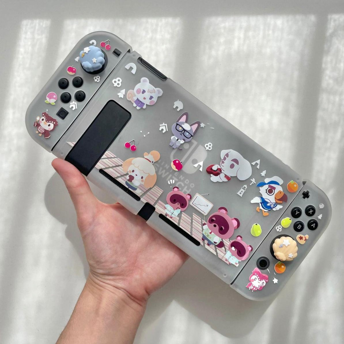A person holds a Nintendo Switch protected by the Chibi Animal Crossing Case from petimint, featuring colorful cartoon animal designs with bears and rabbits under a transparent, soft TPU cover. Sunlight creates gentle shadows in the background.