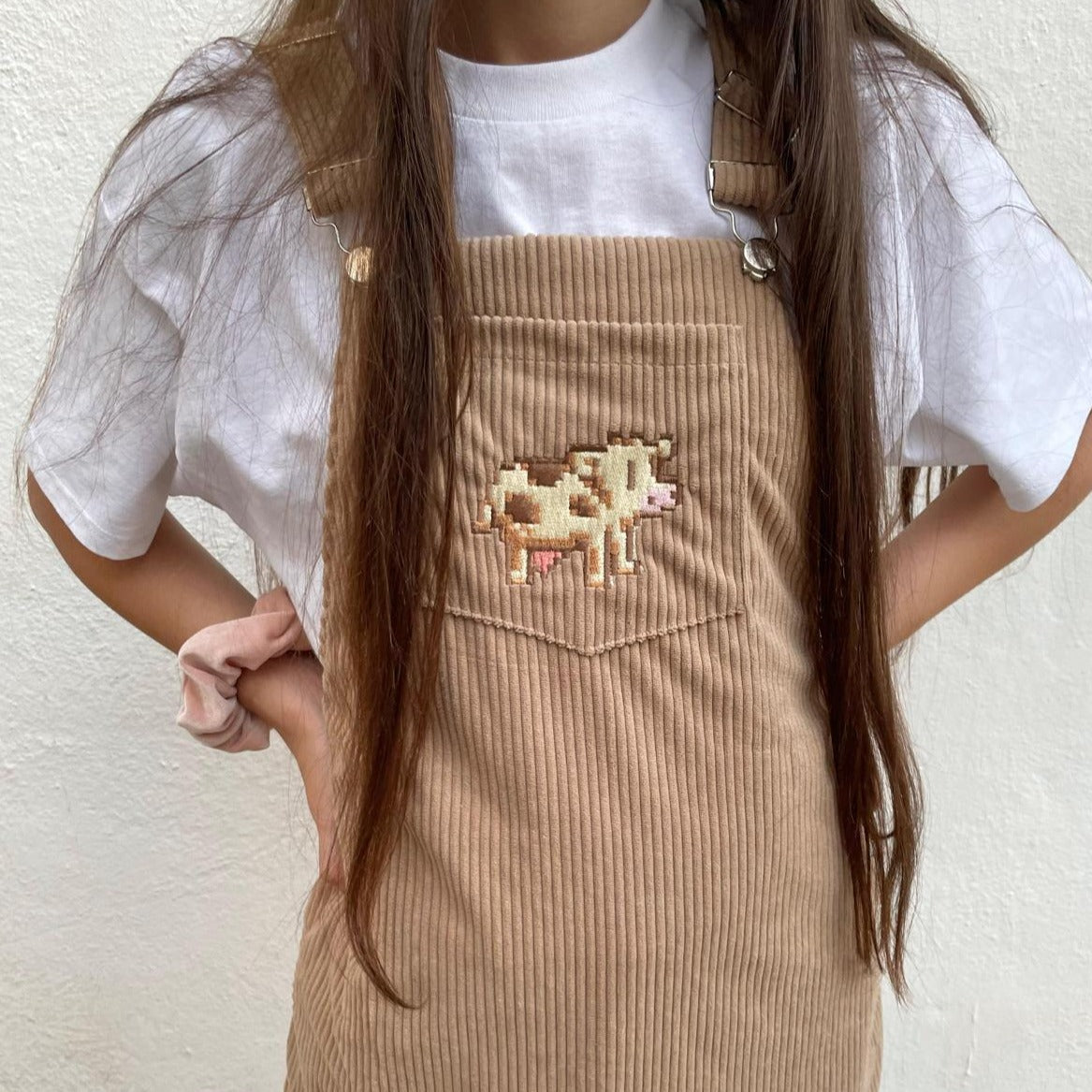 A person wearing petimint's Stardew Cow Overalls Dress, featuring a pixel art cow patch on the pocket. They also have long brown hair and are wearing a white t-shirt underneath the overalls, holding a Nintendo Switch. The background is a plain white wall.