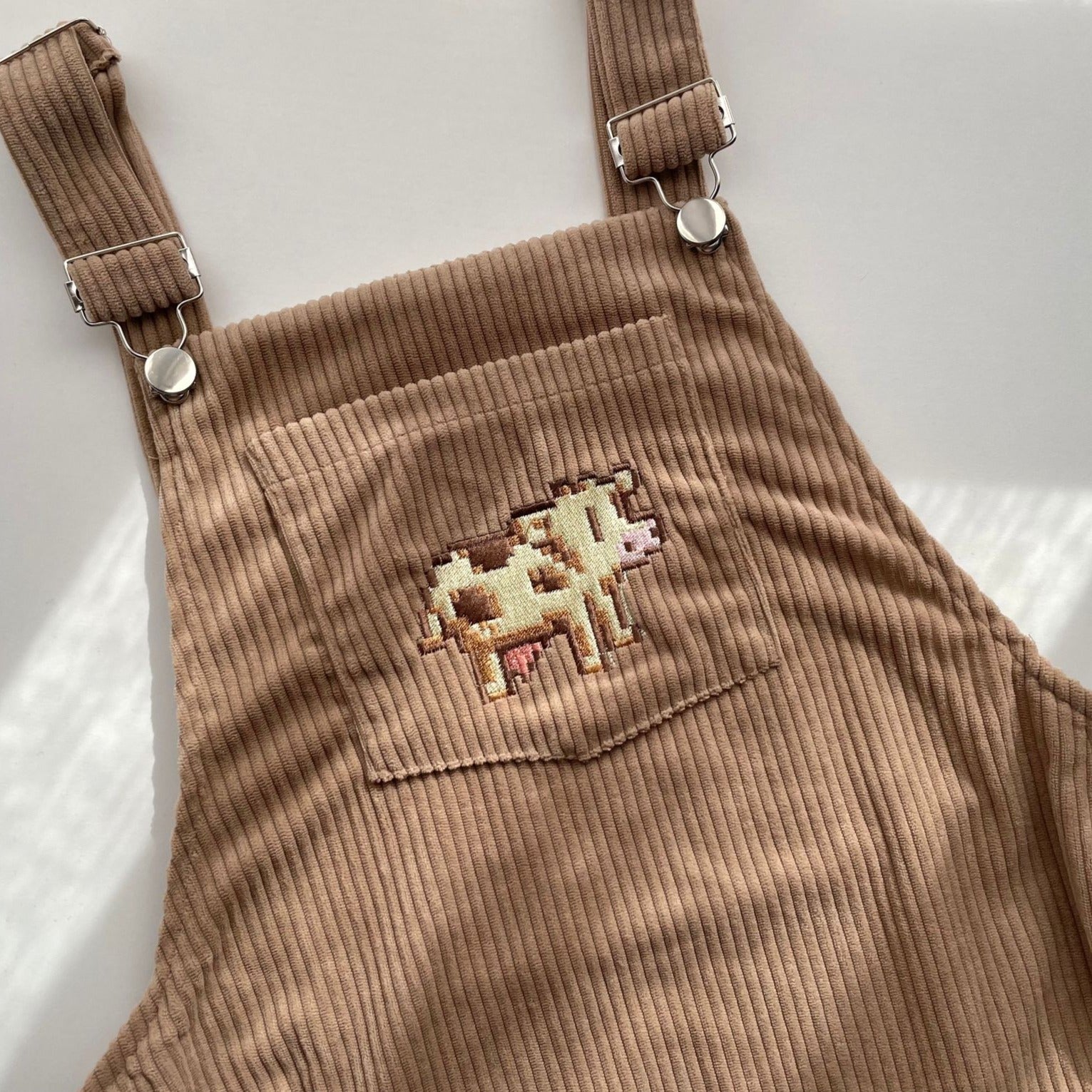 The petimint Stardew Cow Overalls Dress is a pair of beige corduroy overalls featuring silver buckles on the straps. The front pocket showcases an embroidered design of a small, pixel-style cow in shades of brown, white, and pink. The fabric has a textured, ribbed appearance—perfect for the cozygamer who loves their Nintendo Switch.
