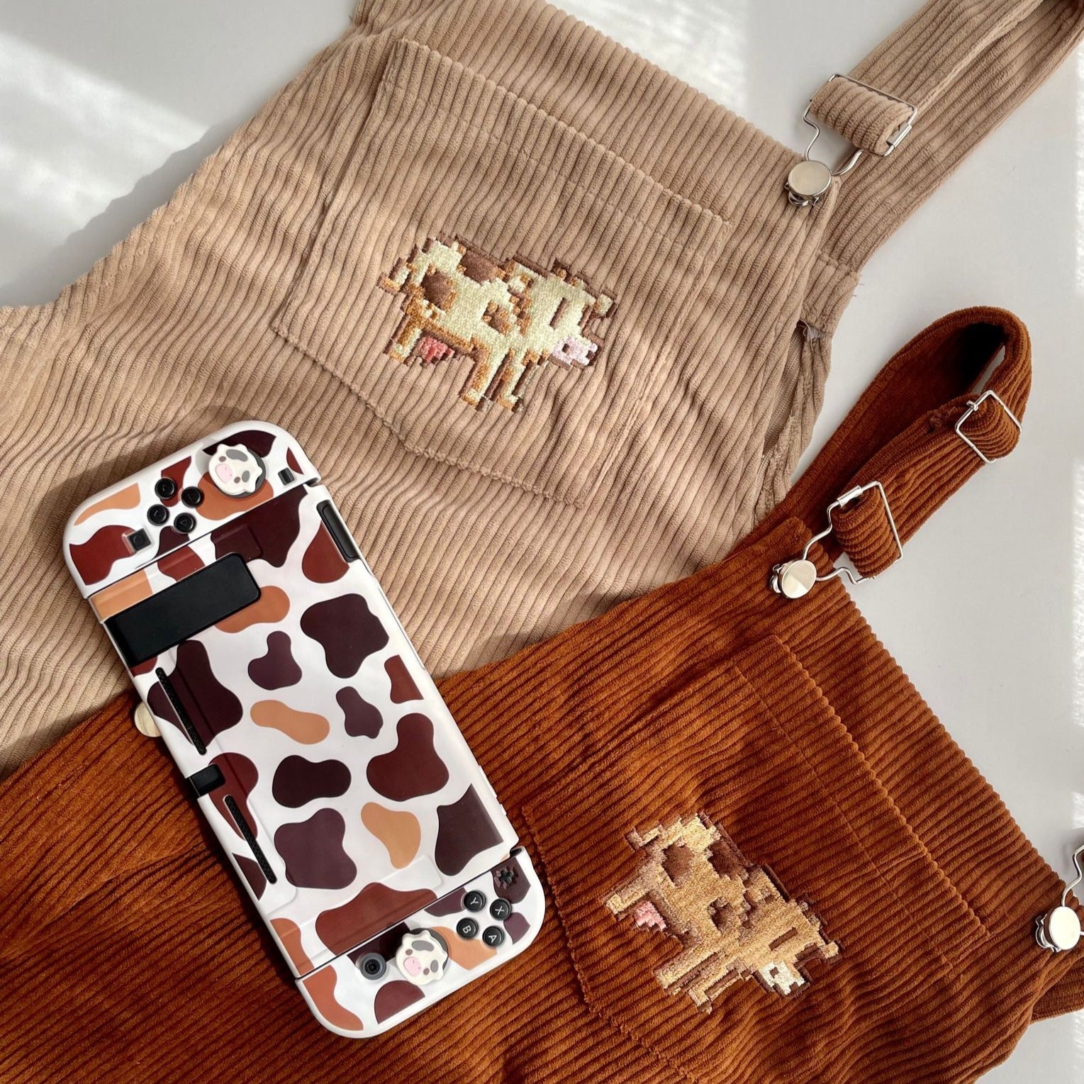Two Stardew Brown Cow Overalls Dresses by petimint, one in beige and one in brown, each featuring an embroidered cow on the chest pocket. Perfect for any gamer, next to them is a handheld gaming console with a cow print case that includes illustrations of a cow’s face. This quirky apparel combination is ideal for gaming enthusiasts!