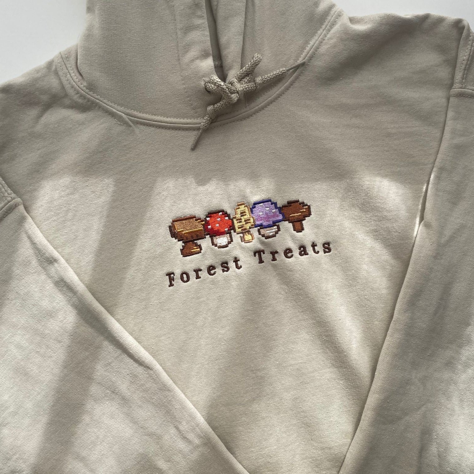The Forest Treats Hoodie by petimint is a beige hoodie with an embroidered design showcasing four colorful mushrooms in a pixel art style, accompanied by the text "Forest Treats" underneath. Ideal for the cozygamer, this hoodie features drawstrings and is pictured laid out flat under natural sunlight, highlighting its fabric.
