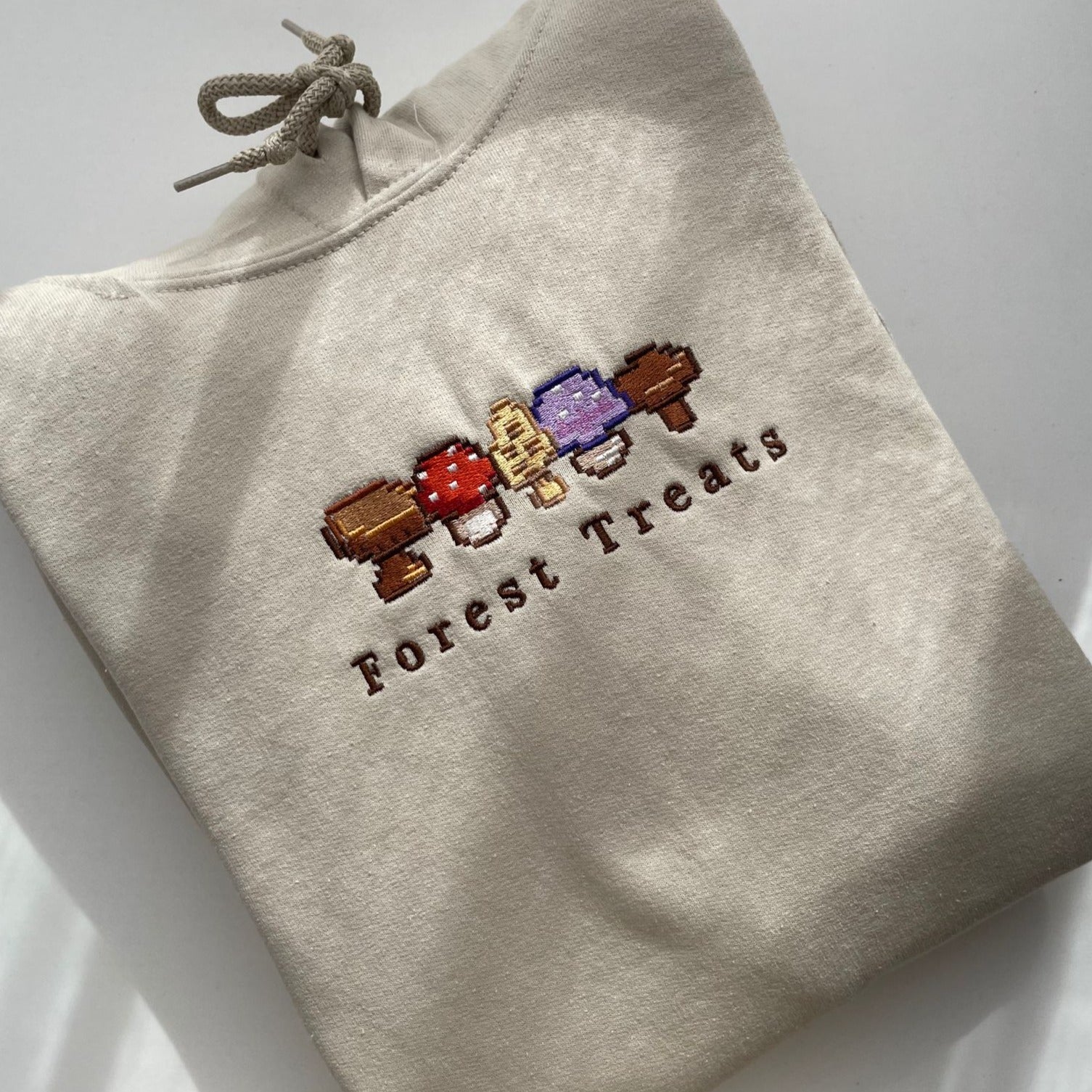 The beige "Forest Treats Hoodie" by petimint, perfect for any cozygamer, is neatly folded and features an embroidered design of five colorful mushrooms. Below the mushrooms, the words "Forest Treats" are embroidered in brown text. This stylish apparel is placed on a white surface with soft lighting.