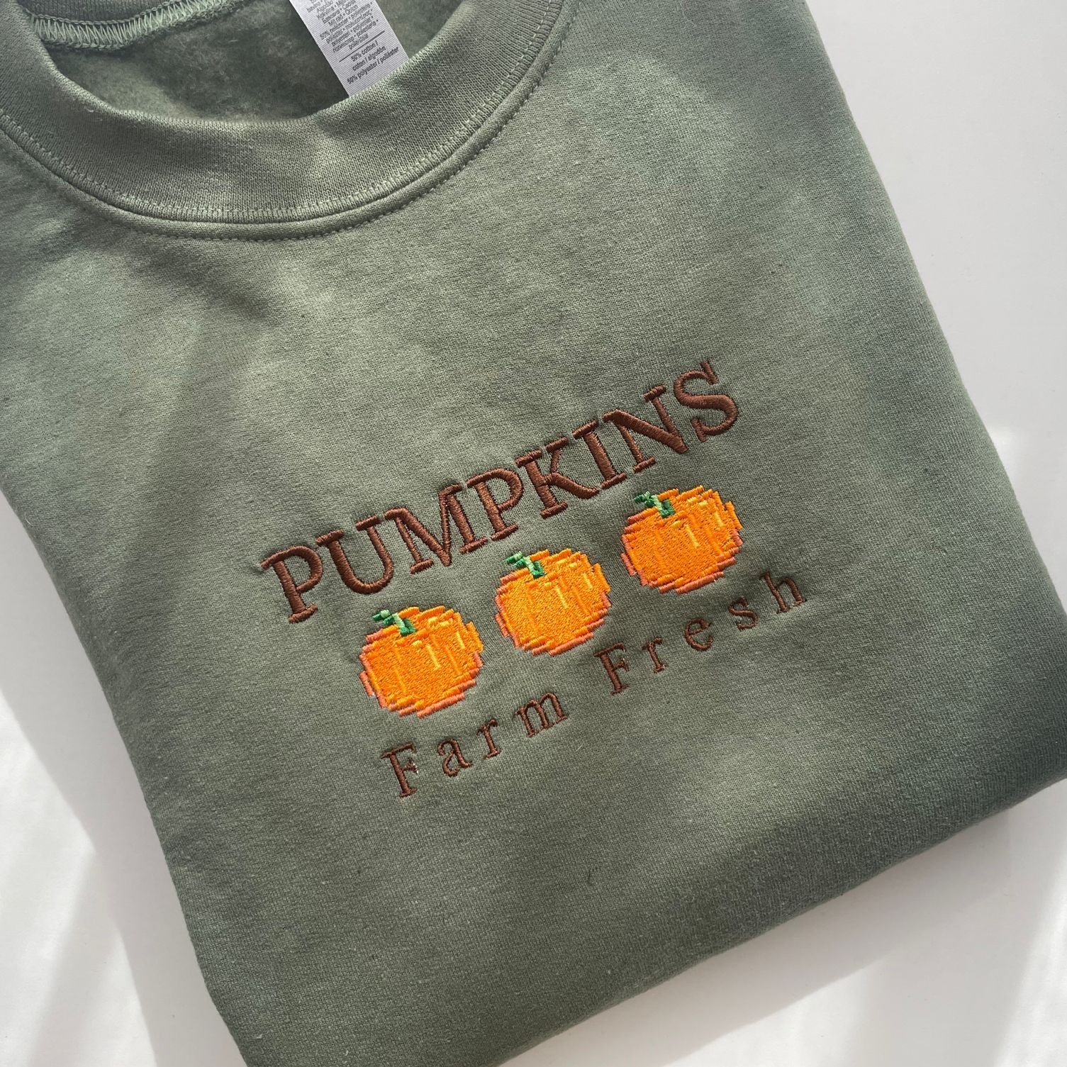 A folded green crewneck sweatshirt from petimint's "Pumpkins Farm Fresh Crewneck Halloween Apparel" collection, featuring an embroidered design on the front. The design showcases the words "PUMPKINS Farm Fresh" with three orange pumpkins in between, all stitched in brown and orange thread. This piece is perfect for any Stardew Valley gamer.