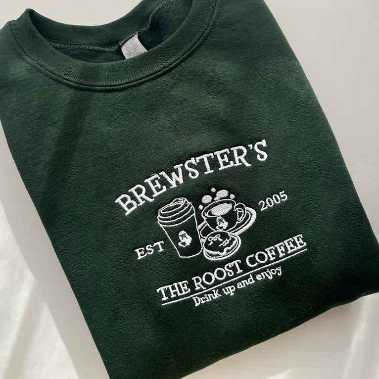 A folded dark green Brewster's Crewneck from petimint, featuring white embroidery on the chest. The text reads "Brewster's EST 2005 THE ROOST COFFEE drink up and enjoy" surrounding images of a takeout coffee cup, a coffee mug, and a plate with a small dessert. Perfect cozy gamer apparel for those gaming marathons.