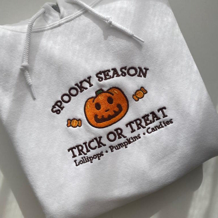 The Jack Spooky Season Hoodie by petimint is displayed with embroidery of a smiling orange pumpkin in the center. Above the pumpkin, text reads "Spooky Season," and below it reads "Trick or Treat." There are also small embroidered images of lollipops and candies around the pumpkin. This white hoodie is perfect apparel for gamers to wear while playing their Nintendo Switch.