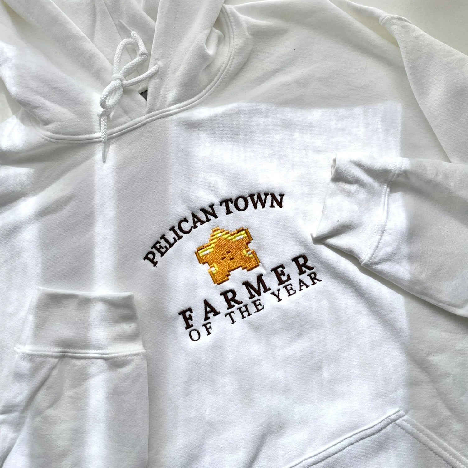 The "Farmer of the Year Hoodie" by Petimint is a white hoodie featuring the text "PELICAN TOWN" in an arc above a pixelated golden trophy. Below the trophy, it reads "FARMER OF THE YEAR." This gamer apparel includes a front pocket and drawstrings at the hood.