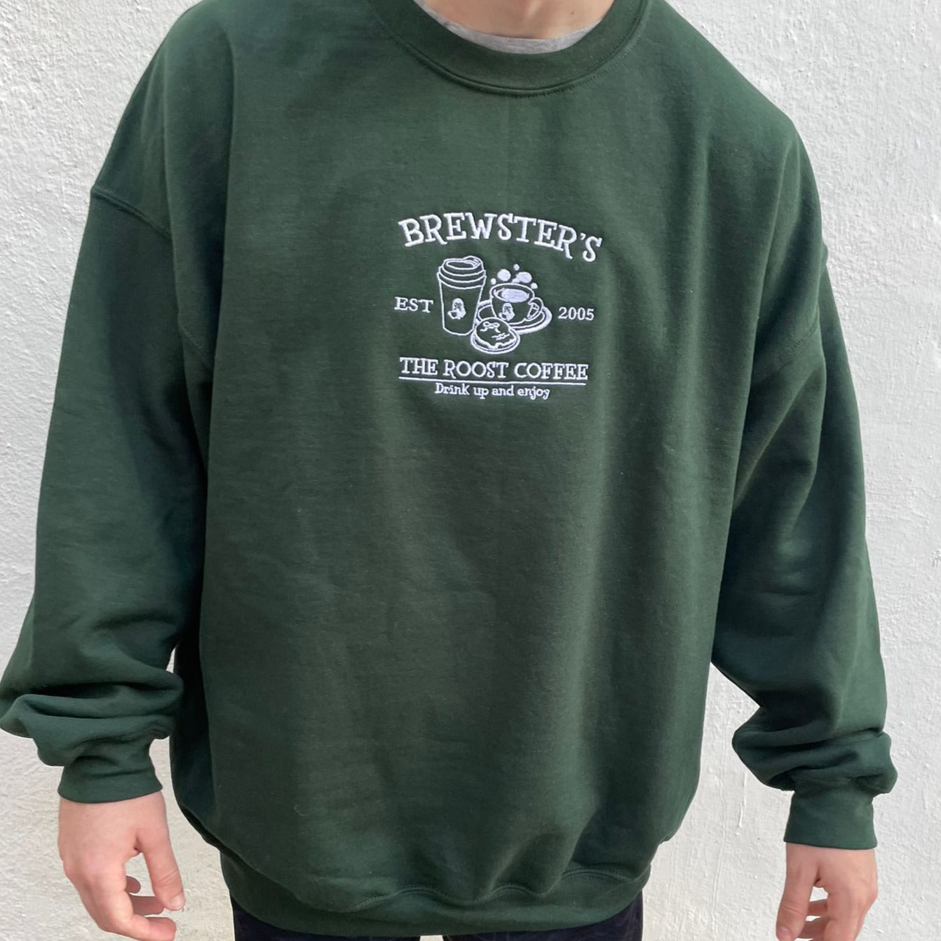 A cozygamer stands against a light-colored wall, clutching their Nintendo Switch and ready for some relaxing gameplay. They're wearing petimint's Brewster's Crewneck, a dark green sweatshirt adorned with the text "BREWSTER'S EST 2005 THE ROOST COFFEE Drink up and enjoy" along with an illustration of a coffee cup, a rooster, and a sunrise.