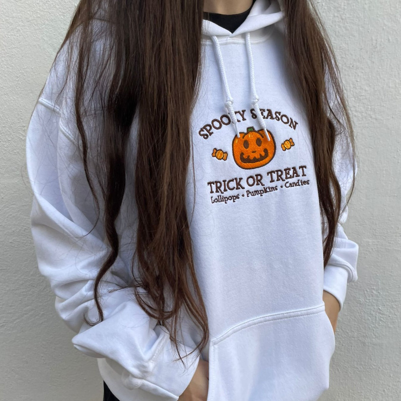 A person with long brown hair wears the petimint Jack Spooky Season Hoodie, which features a pumpkin and text that reads "Spooky Season, Trick or Treat, Lollipops - Pumpkins - Candies." Holding a Nintendo Switch, they strike the perfect cozygamer vibe against a plain white wall.