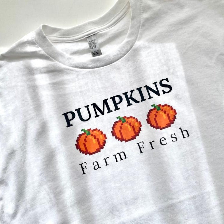 The "Pumpkins farm fresh T-Shirt Halloween Apparel" by petimint features the text "PUMPKINS" in bold black letters above three pixelated orange pumpkins. Below the pumpkins, the text reads "Farm Fresh" in smaller black letters. Perfect for cozy gamers and Halloween lovers, this white t-shirt is laying flat on a smooth surface.