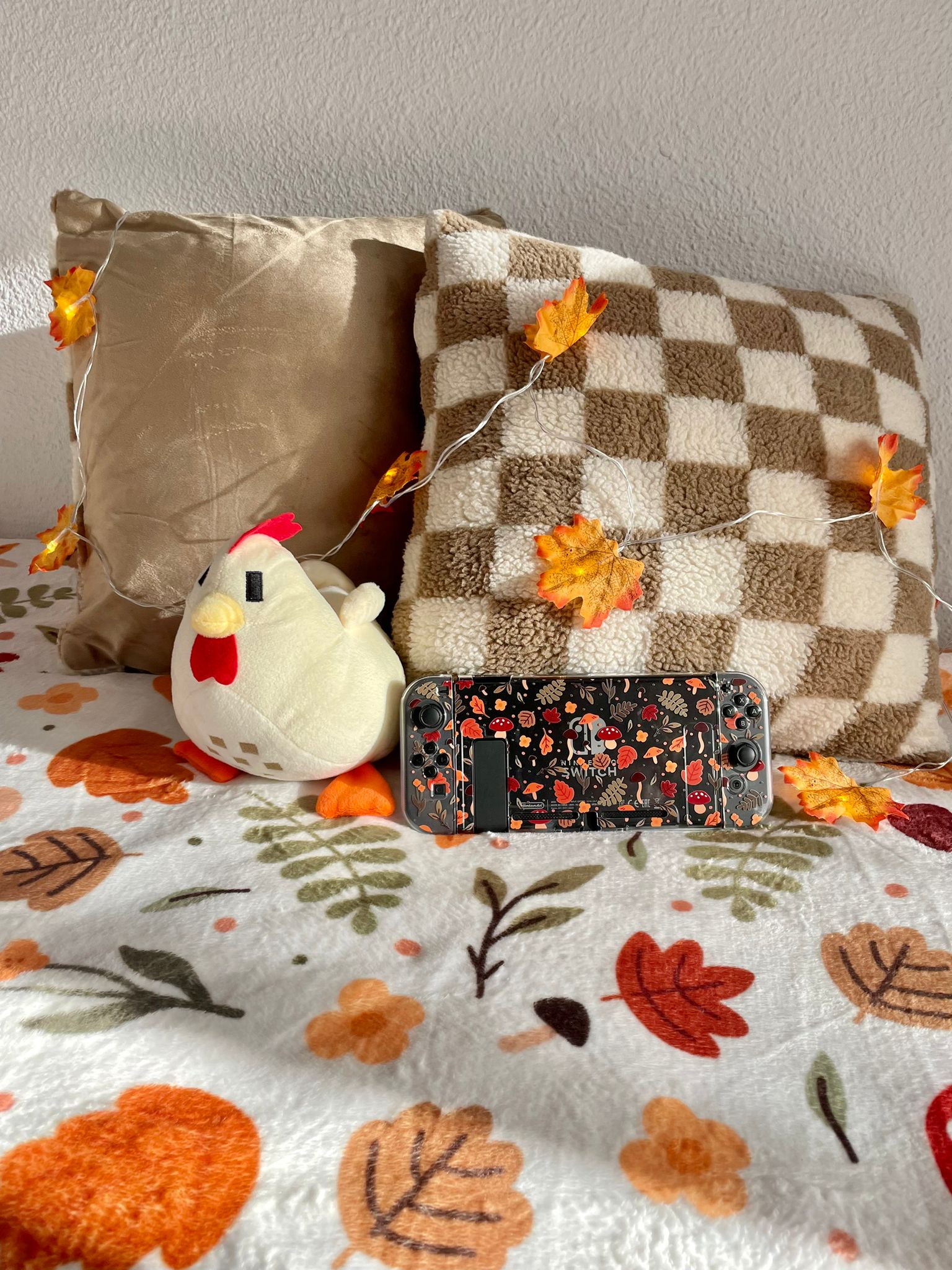 A cozy scene featuring two pillows—one solid beige and one checkered brown and white—adorned with a string of faux autumn leaves. In front, a plush chicken toy and a handheld gaming console with an autumn-themed cover rest on the "Cozy Autumn Soft Blanket" from petimint, enhancing the home decor.
