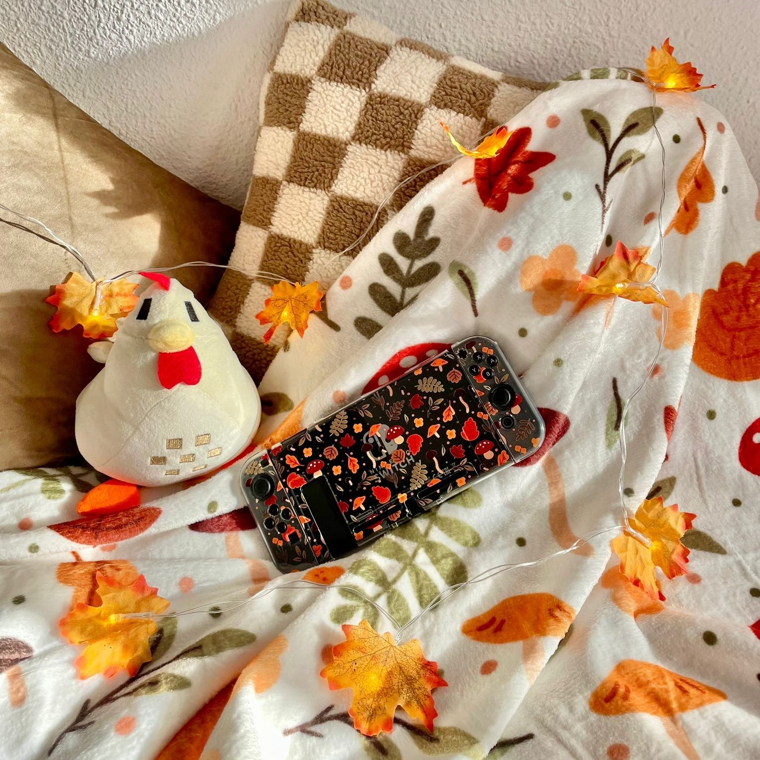 A cozy scene featuring a beige and brown checkered pillow, a plush toy chicken, and a knitted blanket with autumn-themed designs. A handheld gaming console with a black case lies on the blanket surrounded by decorative fall leaves, creating the perfect setup for any cozy gamer.