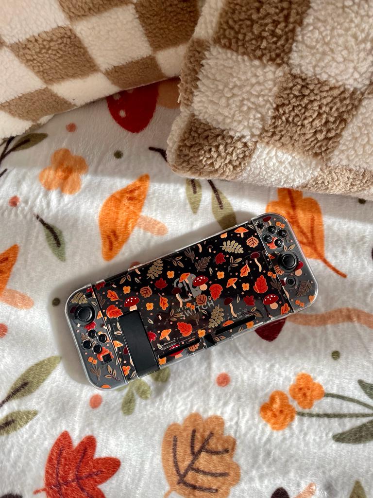 A handheld gaming console with an autumn-themed skin featuring leaves, mushrooms, and acorns rests on a bed adorned with seasonally printed sheets and two blankets—one checkered and the other a Cozy Autumn Soft Blanket by petimint, perfect for enhancing your home decor.