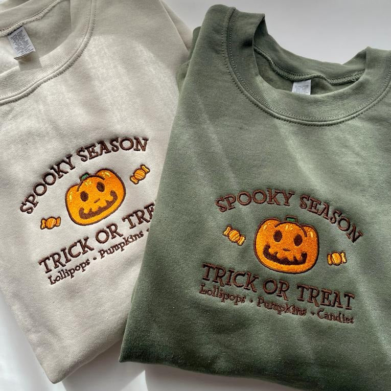 Two crewneck sweatshirts from petimint are displayed side by side, perfect for cozy gamers. Both feature an embroidered design of a smiling jack-o'-lantern, with the text "Spooky Season" on top and "Trick or Treat" below, along with small images of candy. One sweatshirt is off-white, and the other is olive green—essential apparel for fall.