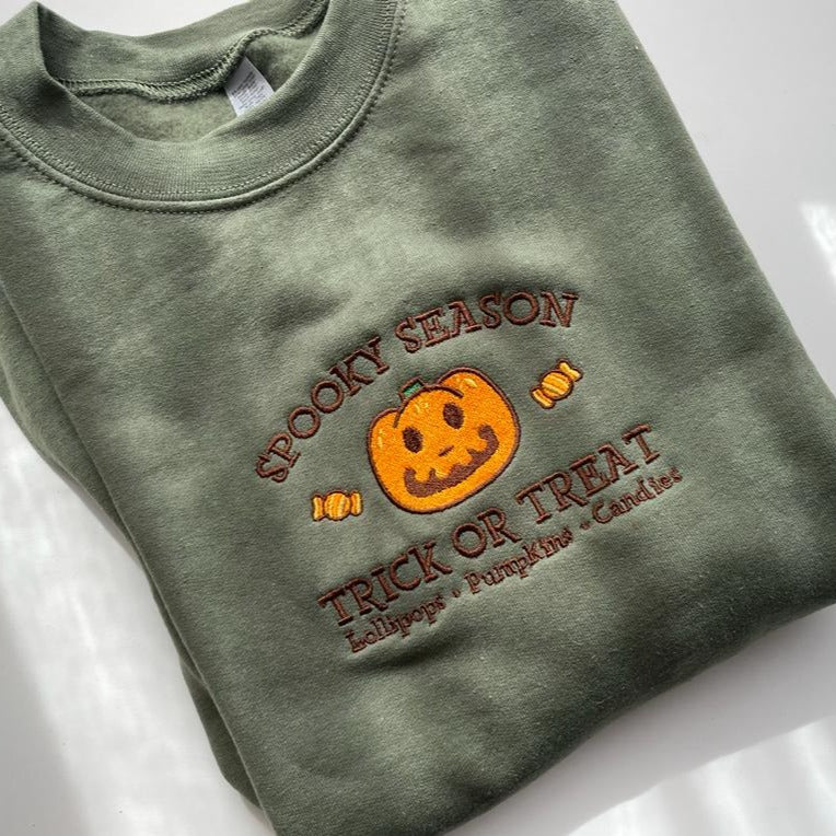 The Jack Spooky Season Crewneck by petimint is a folded green sweatshirt adorned with Halloween-themed embroidery showcasing a cheerful jack-o'-lantern and candy. The text "Spooky Season" appears above the pumpkin, with "Trick or Treat" followed by "Lollipops, Pumpkins, Candies" below. This cozy apparel is ideal for gamers enjoying their Nintendo Switch sessions.