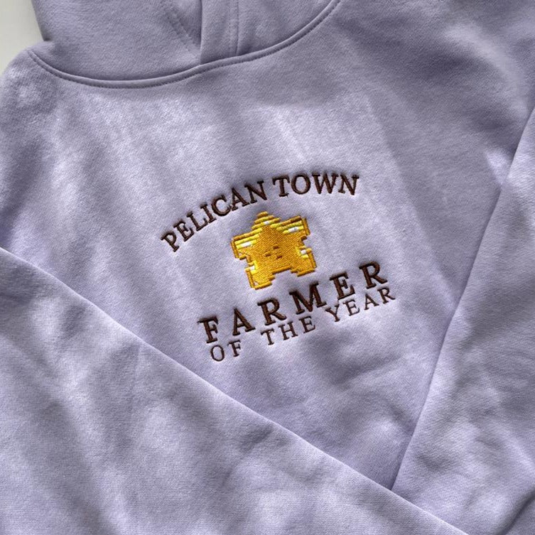 A folded lilac "Farmer of the Year" hoodie from the brand petimint is displayed, featuring an embroidered logo with a golden star and the text "PELICAN TOWN FARMER OF THE YEAR" in brown stitching. This cozy garment is ideal for any gamer looking to show their pride while playing on their Nintendo Switch.