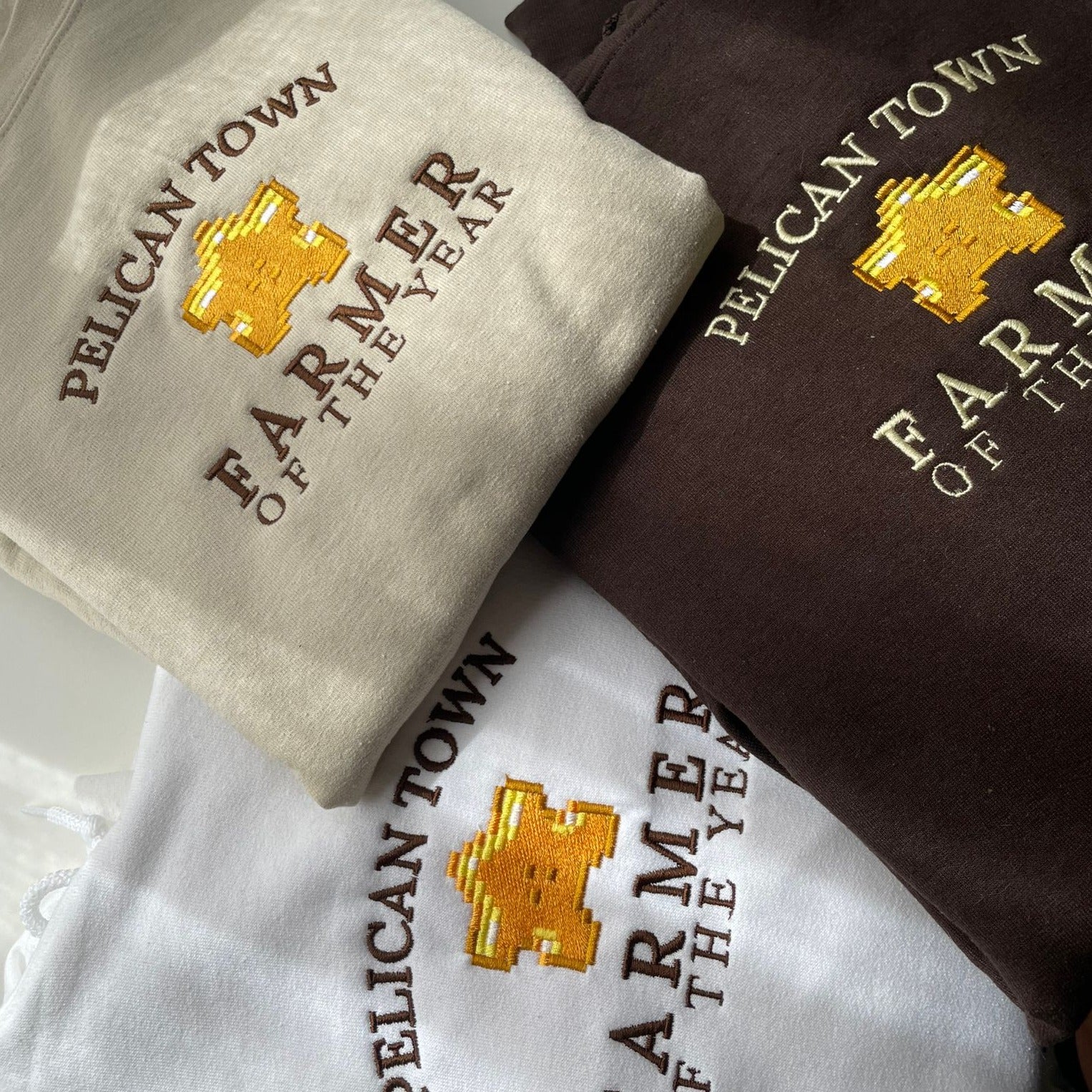 Three folded "Farmer of the Year" hoodies from petimint, in light tan, brown, and white hues. Each hoodie features embroidered text: "PELICAN TOWN FARMER OF THE YEAR" with a golden pixelated trophy at the center. Perfect for any cozy gamer who loves their Nintendo Switch farming games. The hoodies are arranged neatly on a flat surface.