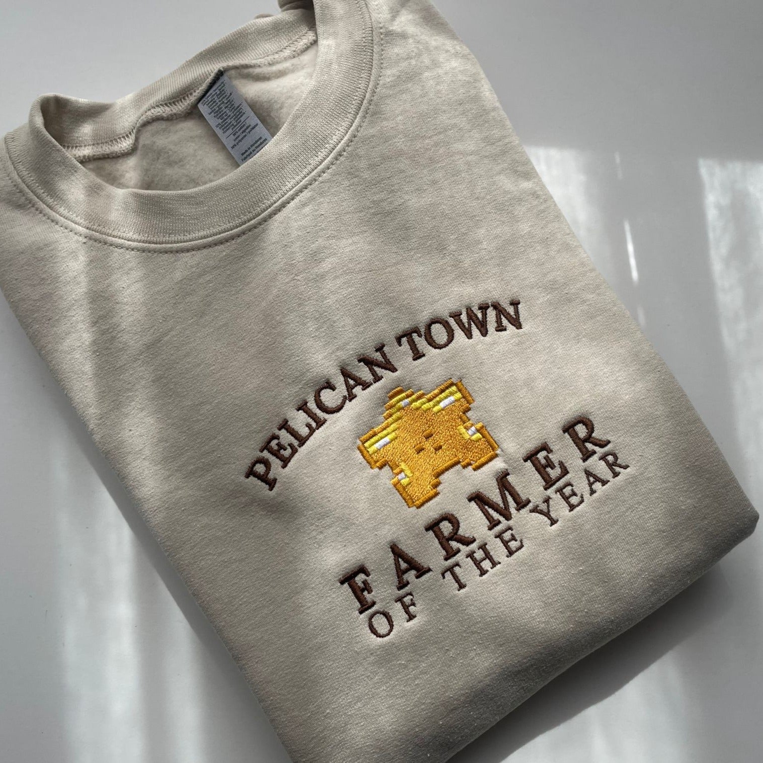A folded beige "Farmer of the Year Crewneck" from petimint features a graphic of a golden pixelated star and the text "PELICAN TOWN FARMER OF THE YEAR" embroidered in dark brown thread on the front. Perfect for cozy gamers, this stylish piece is placed on a smooth white surface, ready to keep you snug during your Nintendo Switch marathons.