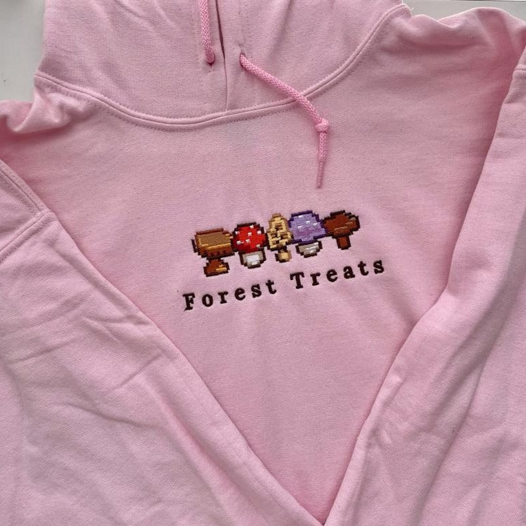 The Forest Treats Hoodie by petimint is a pink hoodie featuring an embroidered pixel art design of various mushrooms in red, brown, purple, and beige arranged in a row above the text "Forest Treats." Perfect for gamers, this hoodie also includes a drawstring hood and can be laid flat.