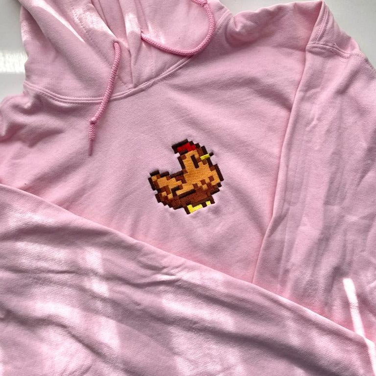 A light pink Stardew Brown Chicken Hoodie by petimint, featuring a pixel art design of a brown chicken on the front, perfect for any gamer. The hoodie is laid out flat, showcasing the design clearly and capturing light and shadow patterns across the fabric, making it an ideal piece of apparel for Nintendo Switch enthusiasts.