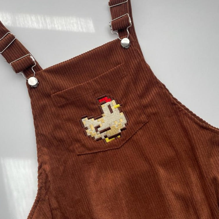 The Stardew Chicken Overalls Dress by petimint is crafted from brown fabric with a ribbed texture and features silver buckle clasps on the straps. The front pocket showcases an embroidered design of a pixelated chicken, making it an ideal choice for cozygamers.