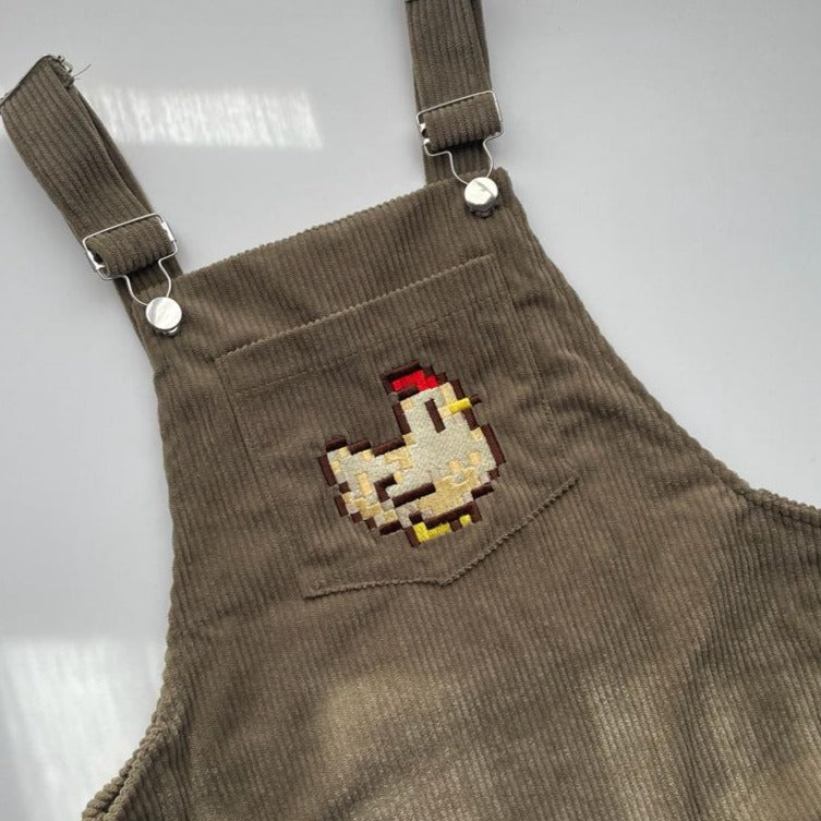 A pair of green Stardew Chicken Overalls Dress by petimint is shown. The front pocket features an embroidered pixel art-style chicken, reminiscent of retro gaming graphics, adding a playful touch to the overall design. Perfect for any gamer looking to showcase their unique style.